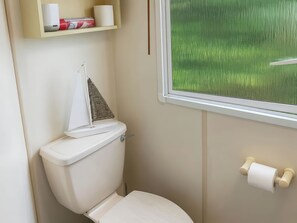 A bathroom with a toilet, a window with a view of green grass, and a shelf with toilet paper and a roll of paper towels. - Brynowen Holiday Park - Holiday Accommodation 19041 (Borth)