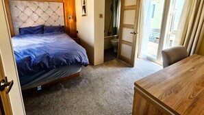 A bedroom with a bed, a wooden dresser, a bathroom with a shower, and a door leading to a balcony. - Ocean Edge Leisure Park - 21286 (Morecambe)