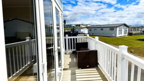 A covered outdoor area with a wooden deck, white railings, and a view of mobile homes in the background. - Ocean Edge Leisure Park - 21286 (Morecambe)