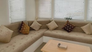Egy nappali, melyben bézs színű, moduláris kanapé, fehér reluxa és egy kis asztal található, melyen egy könyvhalom van.