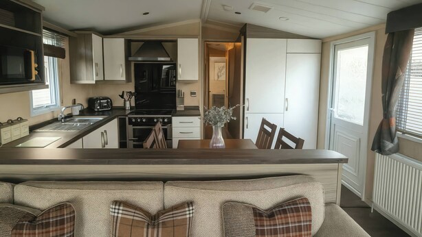 A modern kitchen with a central island, a dining area with a bench, and a view of the adjacent living space. - Whitehouse Leisure Park - 19057 (Abergele)