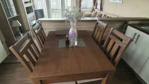 A dining area with a wooden table, chairs, a vase with flowers, and a window with blinds. - Whitehouse Leisure Park - 19057 (Abergele)
