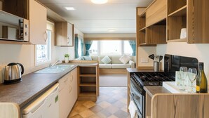 A compact kitchen with a stove, sink, and wooden cabinets. A dining area with a sofa and window is visible in the background.