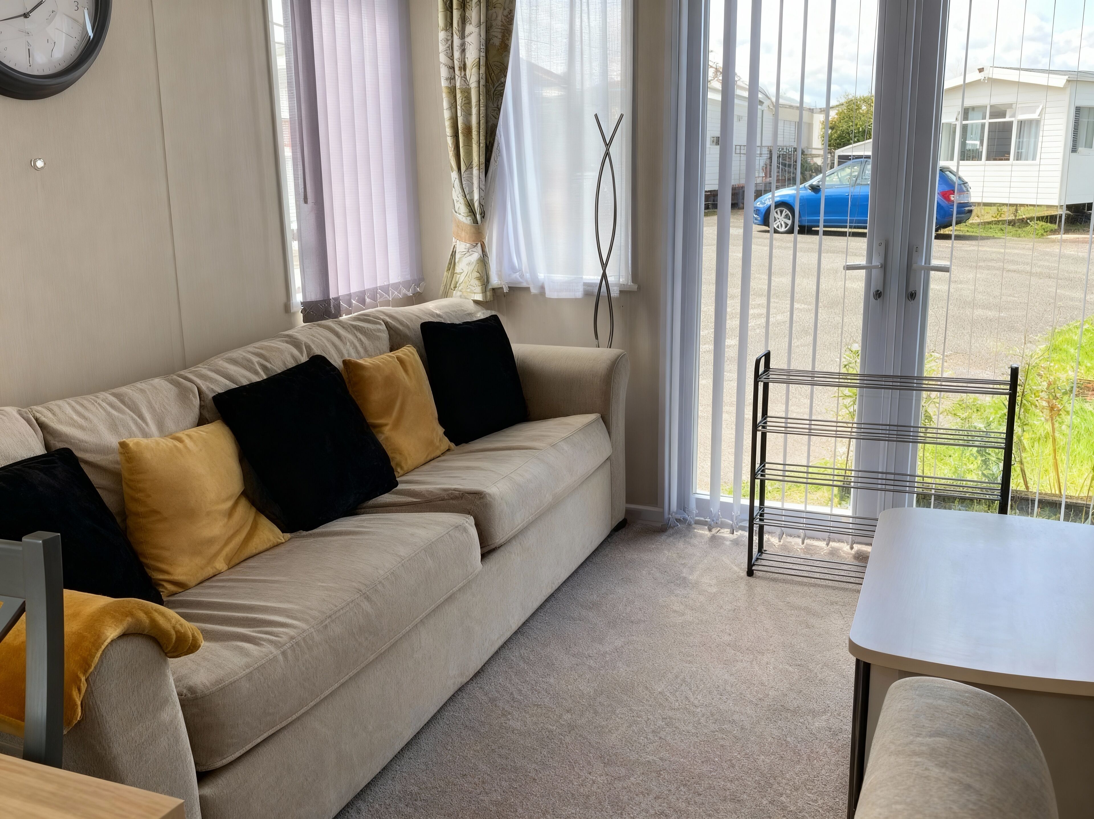 Una habitación con un sofá color beige, cojines negros y amarillos, un reloj en la pared y vistas a un coche azul a través de una ventana.