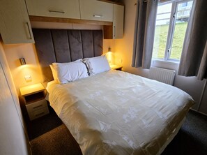 Ein ordentlich bezogenes Bett mit Kopfteil, Nachttischen und einem mit Vorhängen drapierten Fenster.