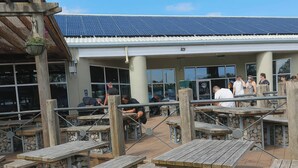 Ein Sitzbereich im Freien mit Steh- und Stehtischen, ein Gebäude mit großen Fenstern im Hintergrund und ein Dach mit Solarpanelen.