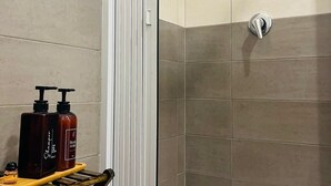 Un espace douche avec une porte blanche, des carreaux de couleur beige et grise, et une étagère en bois où sont rangés les articles de toilette.