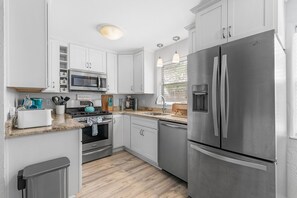A modern kitchen with white cabinets, stainless steel appliances, a granite countertop, and a window with blinds. - Lemon Drop (New Smyrna Beach)