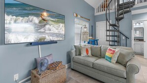 Ein Wohnzimmer mit einem großen Fernseher, der eine Surf-Szene zeigt, einem grauen Sofa mit Surf-Motiv-Kissen, einem Korbwäschesammler und einer Treppe.
