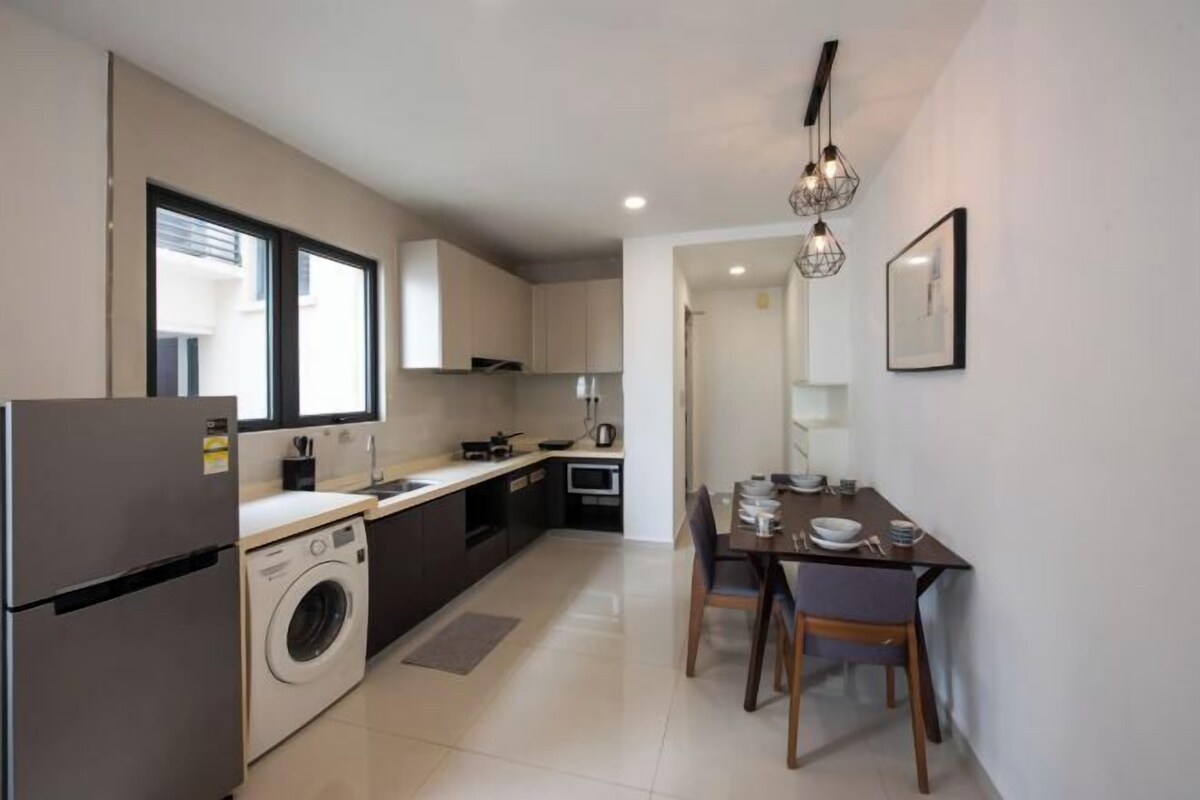 A compact kitchen with a washing machine, a dining area with a table and chairs, and a large window providing natural light.