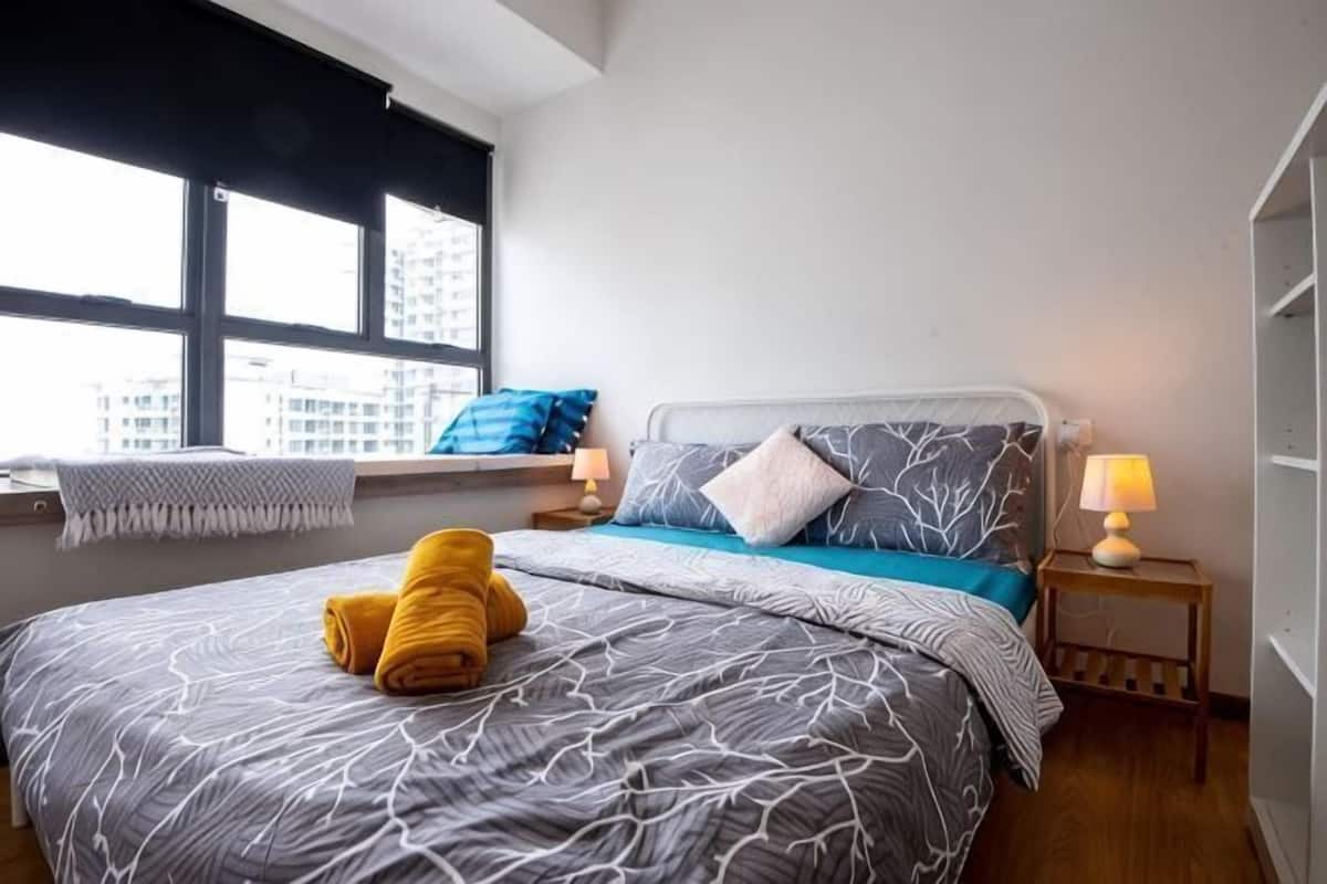 Une chambre avec un grand lit, une fenêtre avec des stores, une table de chevet avec une lampe, et une vue sur les bâtiments à l’extérieur.