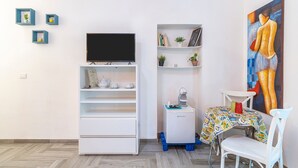 Ein Zimmer mit einer weißen TV-Konsole, einer Kaffeemaschine, einem Tisch mit bunten Tischdecken und einem Stuhl.