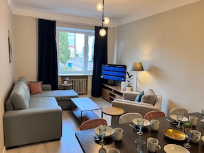 A modern living room with a sofa, coffee table, and dining area set for four. A flat-screen TV is mounted on the wall, and a window provides a view of the outside.