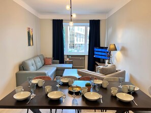 Ein modernes Wohnzimmer mit einer Couch, einem Esstisch mit Tellern und Gläsern, einem Fernseher und einem Fenster mit Vorhängen.