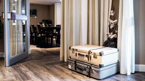 A room with a wooden floor, a large window with blinds, a dining area in the background, and a vintage suitcase in the foreground. - Hotel floor C (Winterberg)