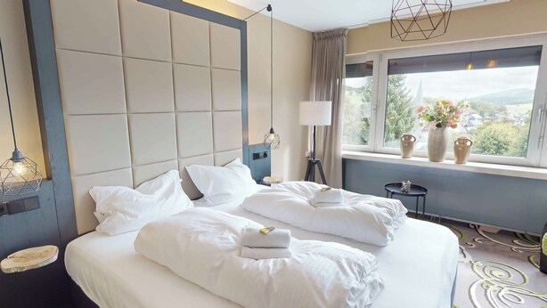 A neatly made bed with white linens and a set of towels, a bedside lamp, and a view of greenery through a window. - Hotel floor C (Winterberg)