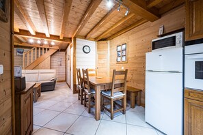 A wooden interior with a dining table, chairs, a refrigerator, and a clock on the wall.