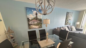 A dining area with a glass table and black chairs, a lamp, and a painting of a lighthouse on the wall.