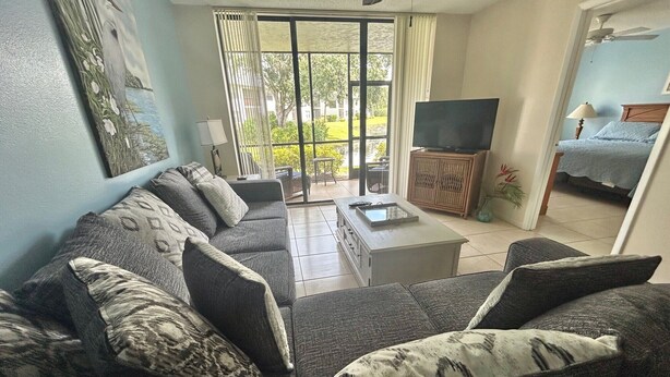 A living room with a sofa, coffee table, and a TV on a stand. There is a sliding glass door leading to a balcony with a view of greenery.