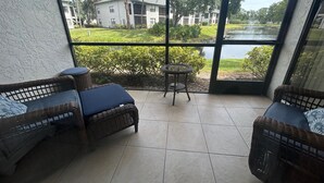 A balcony with wicker furniture and a small table, overlooking a grassy area and a body of water.