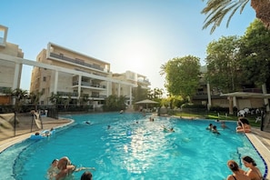 Un área de piscina con varios niveles de edificios, cielo despejado y gente nadando y relajándose.