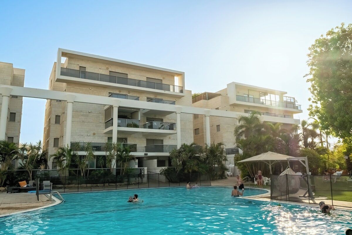 Área de piscina com um edifício em vários níveis ao fundo, cercada por palmeiras e um espaço de estar coberto.