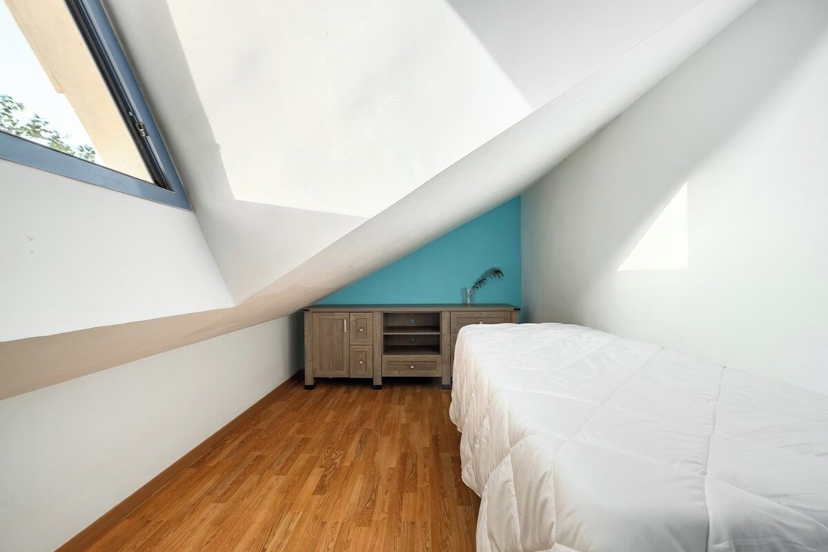 Quarto com teto inclinado, cama, piso de madeira, parede azul e uma janela.