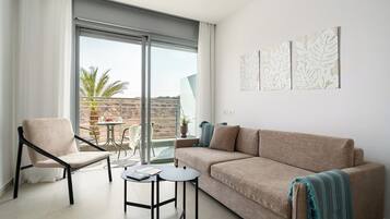 Ein modernes Wohnzimmer mit einer Couch, einem Sessel und kleinen Tischen, einem Balkon mit Aussicht und Kunstwerken an der Wand.