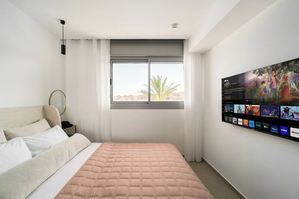 Ein Hotelzimmer mit Bett, großem Fenster mit Vorhängen, einem an der Wand montierten Fernseher, der verschiedene Sender anzeigt, und Blick auf eine Palme.