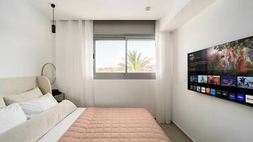 Ein Hotelzimmer mit Bett, großem Fenster mit Vorhängen, einem an der Wand montierten Fernseher, der verschiedene Sender anzeigt, und Blick auf eine Palme.