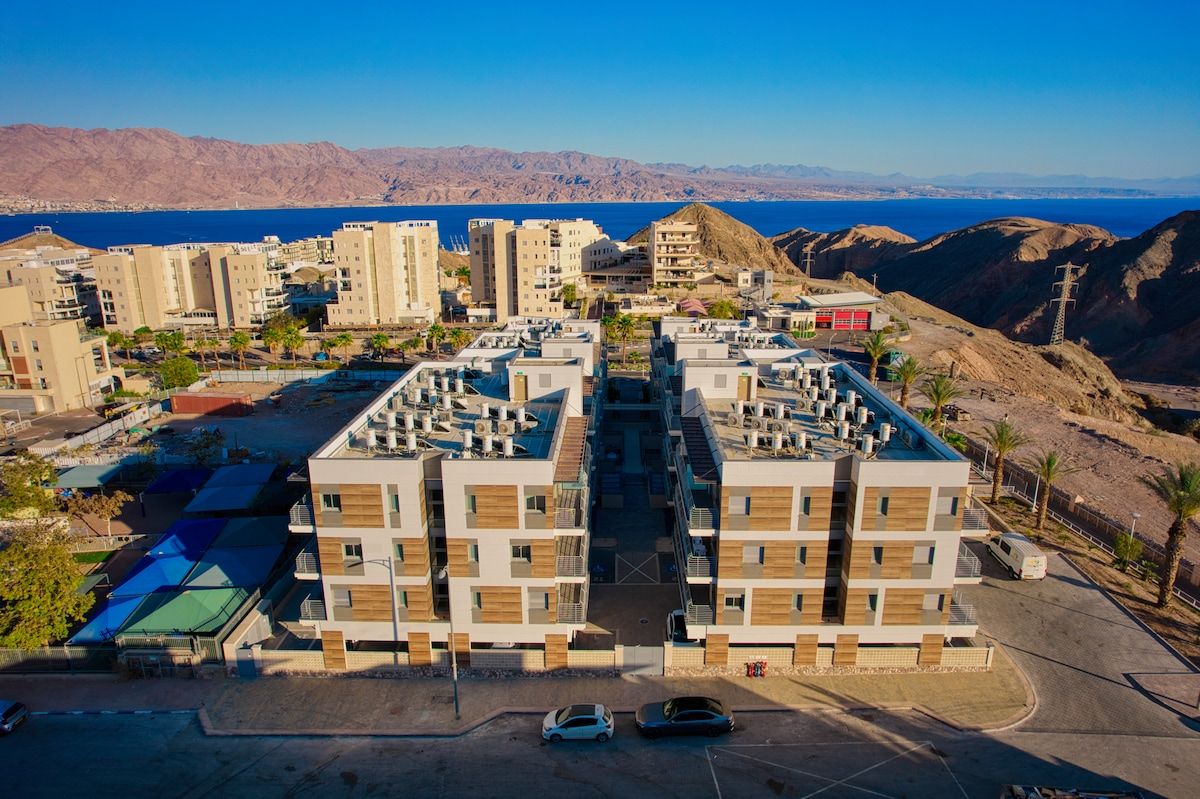 Un immeuble d’appartements moderne doté d’une cour intérieure, entouré de collines désertiques et offrant une vue sur la mer.