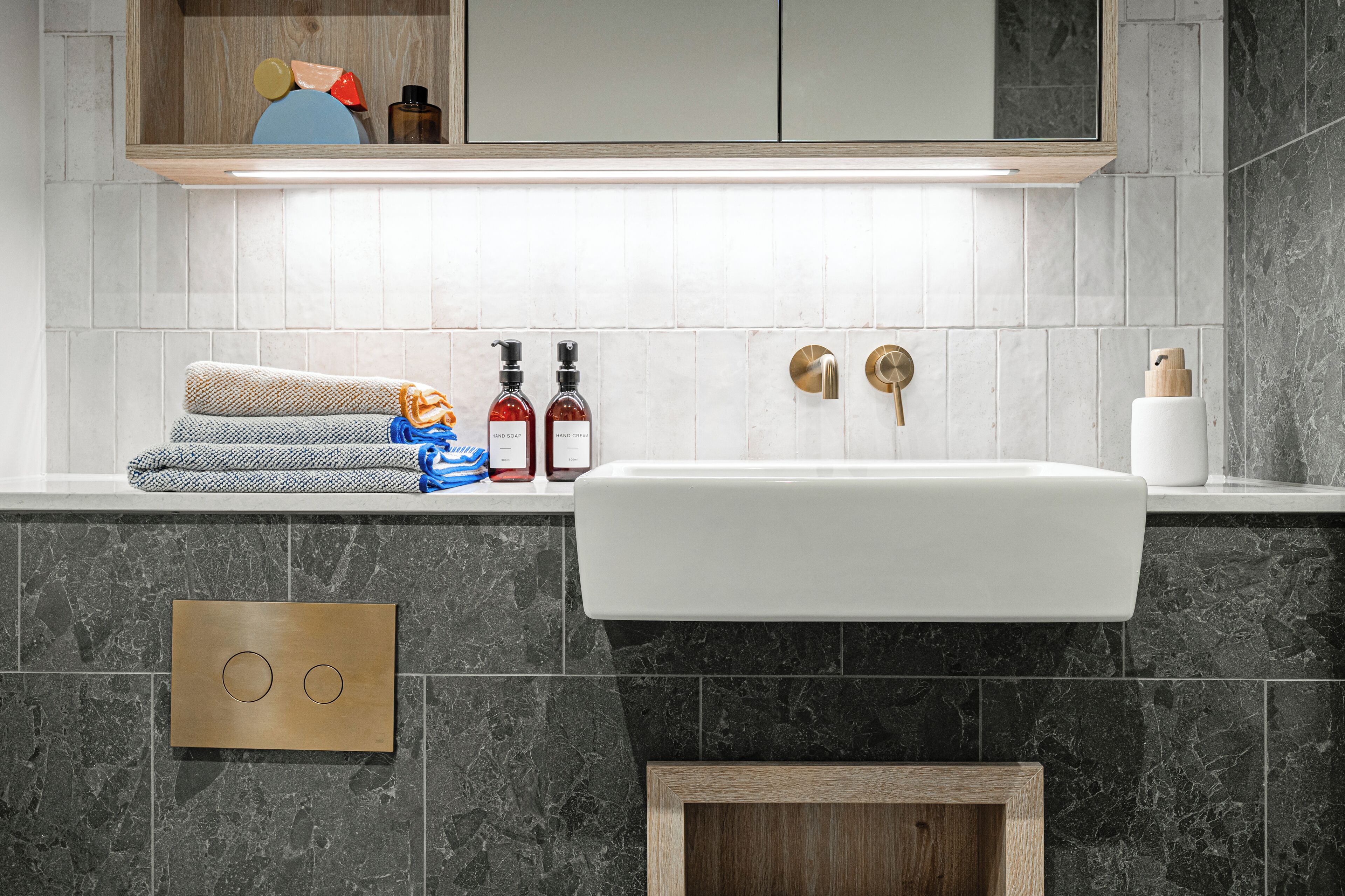 A modern bathroom with a white sink, dark marble tiles, a towel rack with folded towels, and a shelf with toiletries.