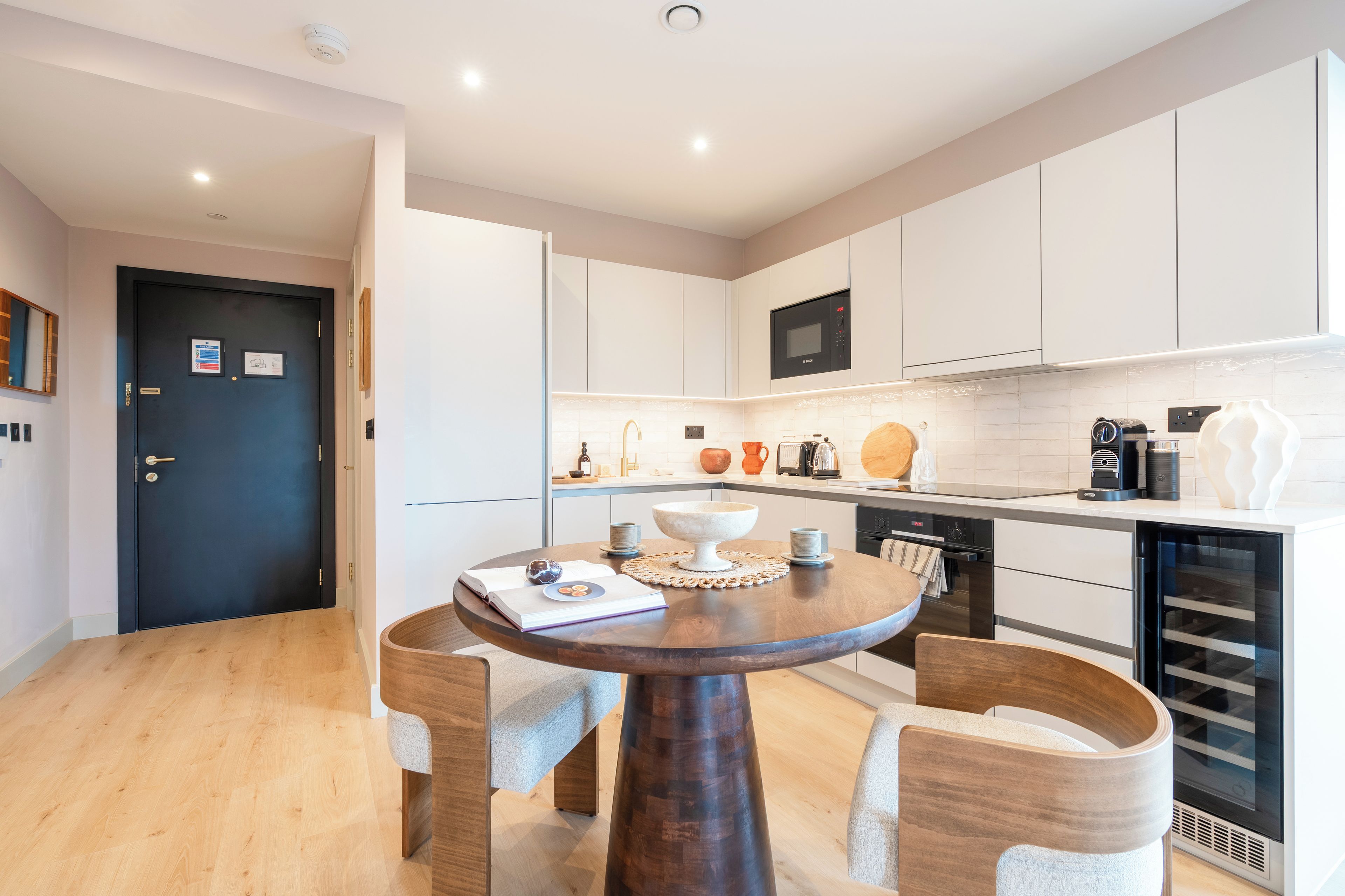 Una cocina moderna con armarios blancos, un microondas negro y un horno empotrado. Hay una mesa de comedor redonda con sillas y una pequeña isla con un tazón y tazas.