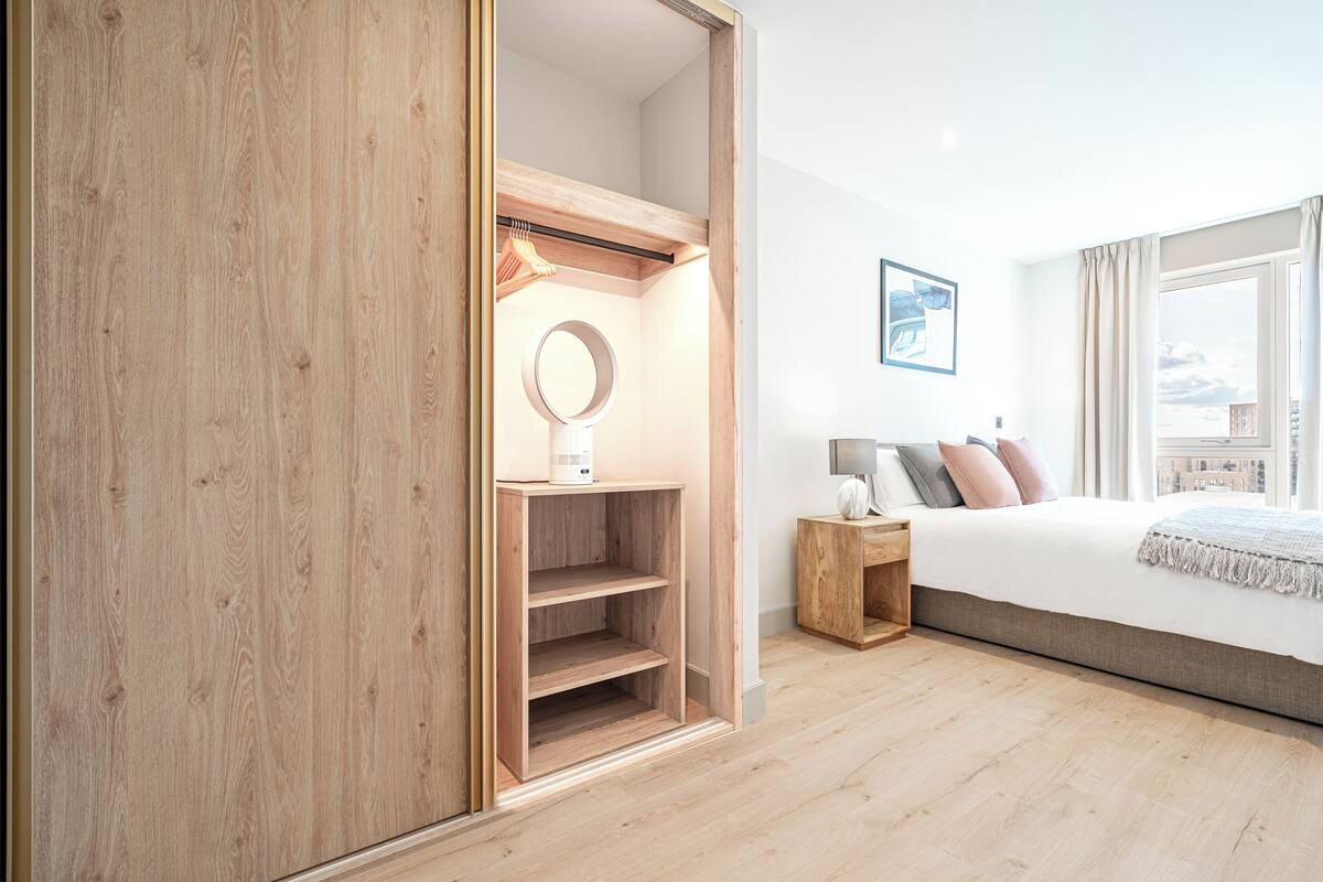 A bedroom with a wooden wardrobe, a bedside table with a lamp, a bed with pillows, and a large window with curtains.