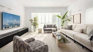 En moderne stue med sofa, lænestol og sofabord, et stort fladskærms-tv og et vindue med lette gardiner.