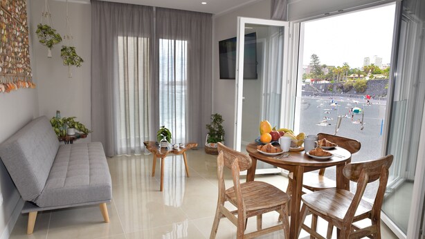 Una sala de estar moderna con un sofá gris, sillas de madera, una mesa redonda con frutas y tazas, y un balcón con vistas a la playa.