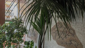Une pièce avec une grande plante en pot, un mur recouvert de carreaux formant une mosaïque, et une vue sur un bâtiment à l’extérieur.