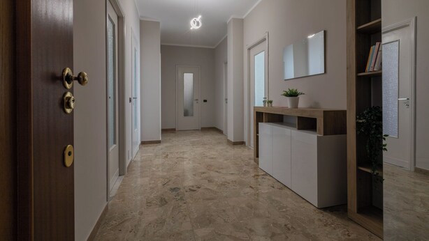 Un couloir doté d’un sol en marbre, d’une porte en bois avec des poignées en laiton, d’une console blanche ornée d’une plante et d’un miroir accroché au mur.