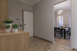 Un couloir avec un comptoir d’accueil en bois, des plantes en pot, une porte et une vue sur un espace repas comprenant une table et des chaises.