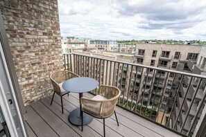 A balcony with two wicker chairs and a small round table, overlooking a cityscape with multiple buildings and a cloudy sky. - Chic Urban Escape in London (London)
