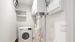 A compact laundry room with a front-loading washing machine, built-in shelving, and visible electrical outlets. - Stylish One-Bed Retreat (London)