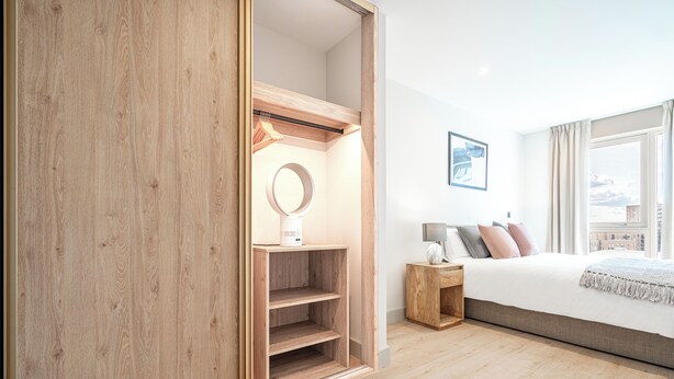 A modern bedroom with a wooden wardrobe, a bedside table with a lamp, a bed with multiple pillows, and a large window with a view of the city. - Stylish One-Bed Retreat (London)