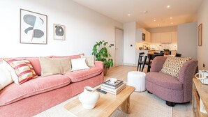 A modern living room with a pink sofa, a purple armchair, and a coffee table. There is a kitchen in the background with white cabinets and a dining area. - Light and Space Retreat (London)