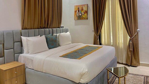 A modern hotel room with a large bed, a bedside table, and a small side table. - The Olive Place (Jos)