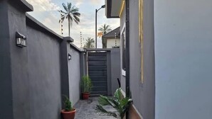 A narrow, paved alleyway with potted plants, a gated entrance, and a building with a grey exterior and wooden accents. - The Olive Place (Jos)