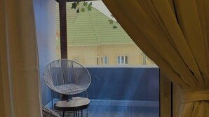 A balcony with a wicker chair and a small table, a view of a building with a green roof, and a curtain partially drawn. - The Olive Place (Jos)