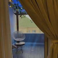 Uma varanda com uma cadeira de vime e uma pequena mesa, vista para um edifício com telhado verde, e uma cortina entreaberta.