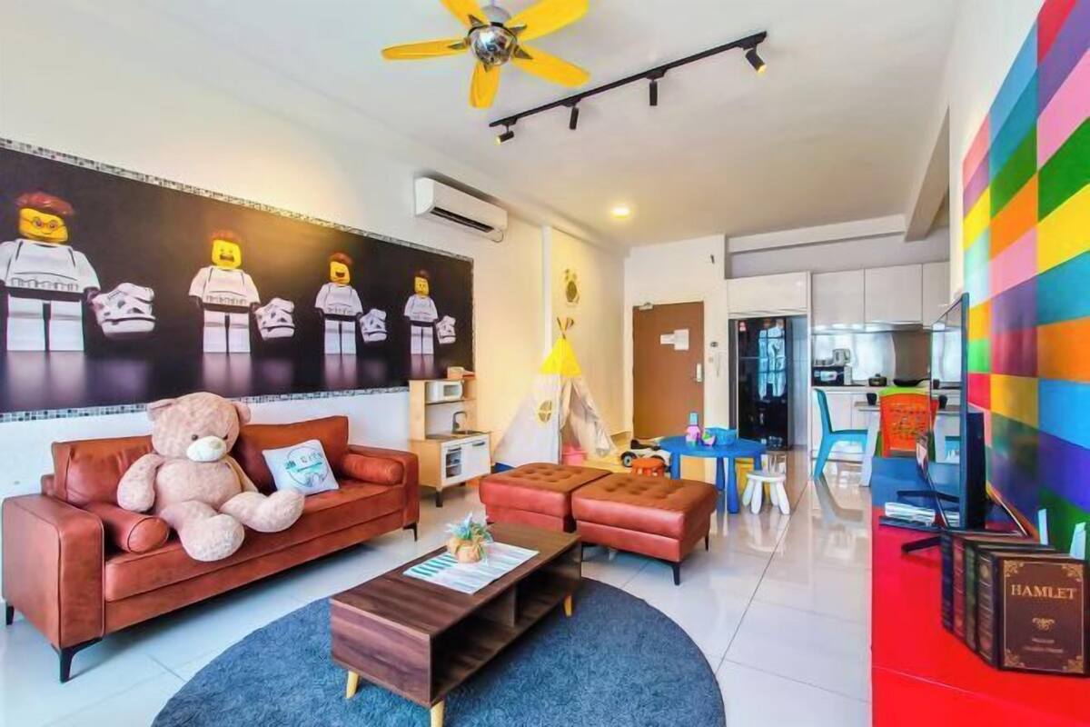 Una habitación de hotel moderna con sofá, una mesita de café, un osito de peluche, una pared con un mural de LEGO, un ventilador de techo y una pared colorida.
