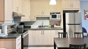 A modern kitchen with light wood cabinets, a stainless steel refrigerator, a microwave, a toaster, and a hanging pendant light. - Central 2BR | Near Metro & Parks (Montréal)
