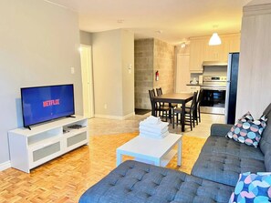 Una sala de estar moderna con un televisor de pantalla plana mostrando 'NETFLIX', una zona de comedor con una mesa y sillas de madera, y una kitchenette al fondo.